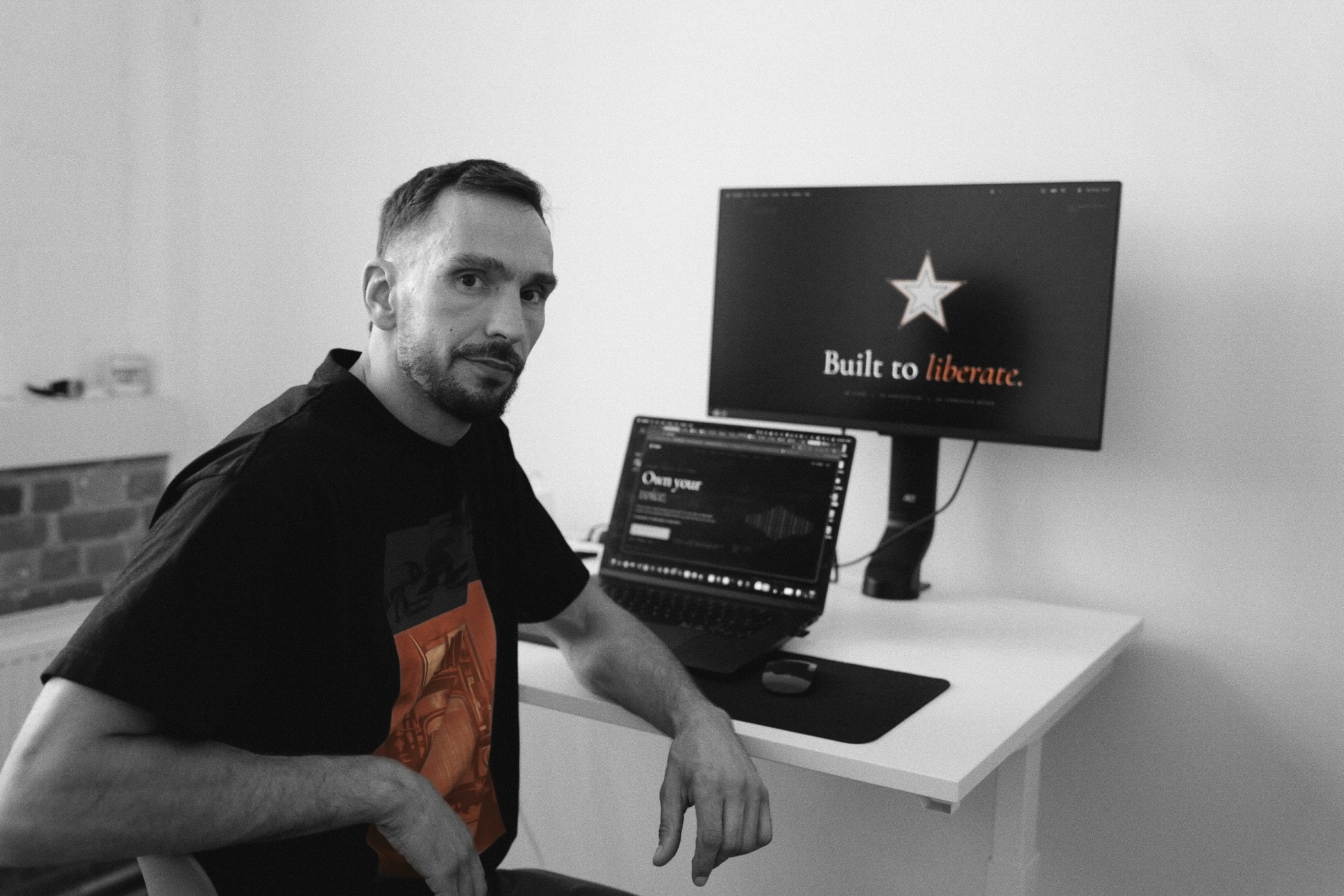 Mehdi at his studio desk, photographed in black and white with red accents on his shirt and screen